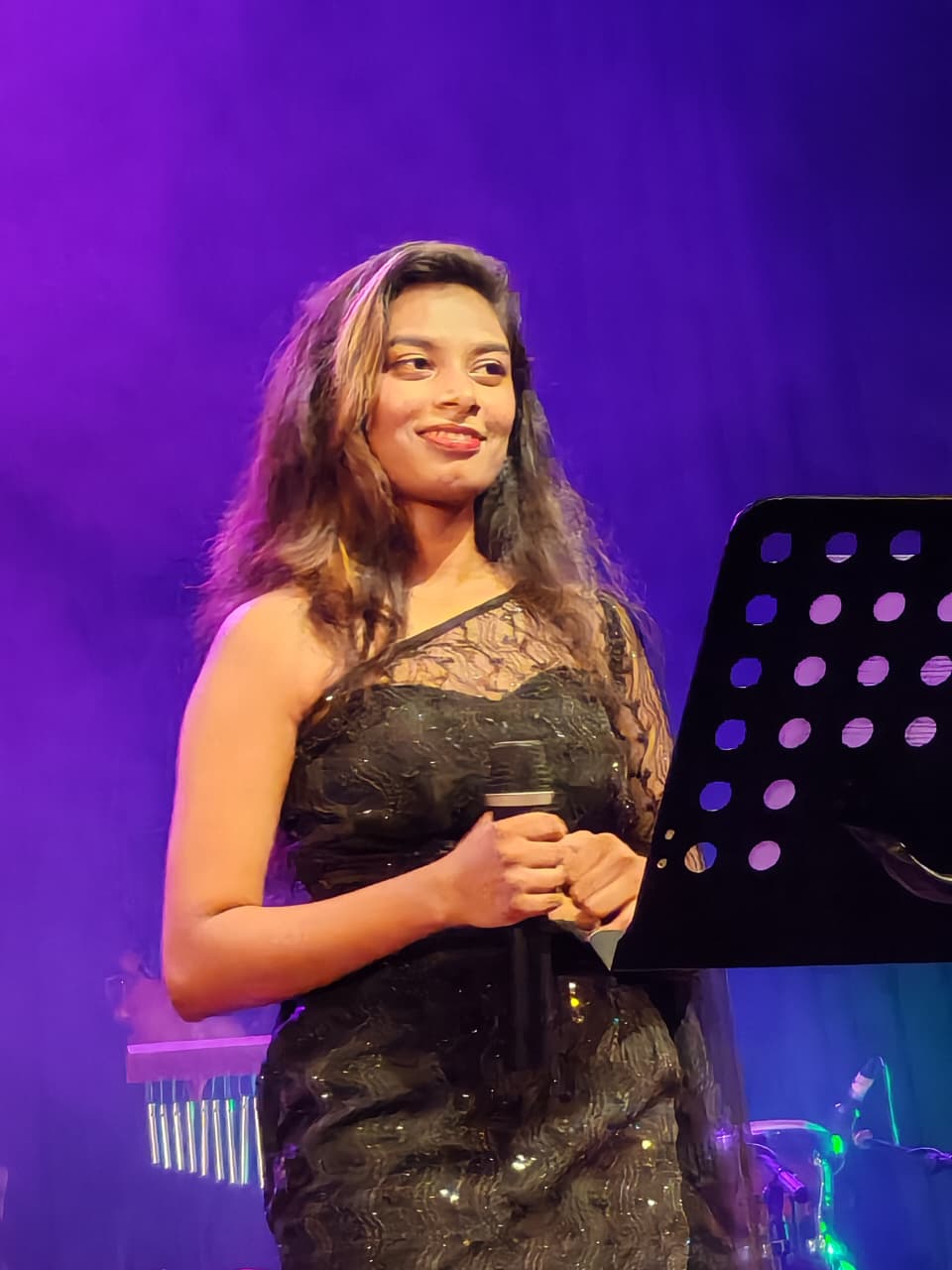Kawya Erandi smiling on stage in black lace gown under purple light